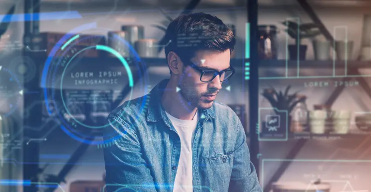 A man wearing glasses and a denim shirt works on a computer, surrounded by futuristic digital interface graphics, in a modern room with shelves and plants in the background.