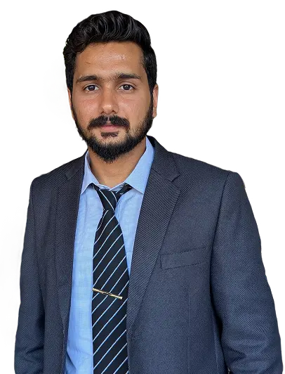 A man with dark hair and a beard wearing a grey suit, light blue shirt, and striped tie, standing and looking at the camera with a neutral expression.