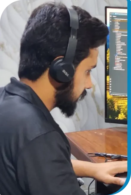 A man with a beard wearing headphones is sitting at a desk, using a computer with code visible on the monitor.